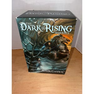 May include: A rectangular book box set titled The Dark Is Rising Sequence by Susan Cooper. The cover features a dark, atmospheric illustration of a hooded figure riding a horse through a forest, with a smaller figure standing in the foreground.