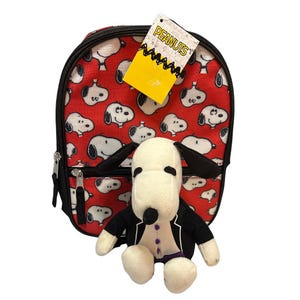 Może przedstawiać: Czerwony plecak ze wzorem Snoopy'ego i małą czarną kieszenią na zamek. Pluszowa zabawka Snoopy w smokingu znajduje się przed plecakiem. Do plecaka przymocowana jest żółta etykieta z napisem "PEANUTS".