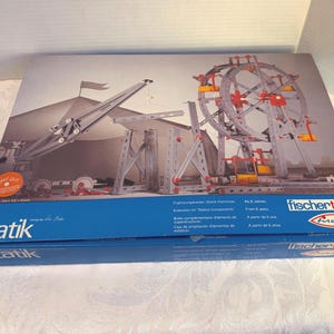 Puede incluir: Una caja del juego de construcción fischertechnik Statik. La caja presenta una ilustración de una grúa, una noria y otras estructuras. La caja es azul con texto blanco y el logotipo de fischertechnik.
