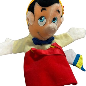 以下が含まれることがあります： 黒い髪、黄色い帽子、青い目のぬいぐるみピノキオの人形。人形は黄色のシャツ、青い蝶ネクタイ、赤い帽子を着ています。腕は広げられています。クラシックな子供のおもちゃ。