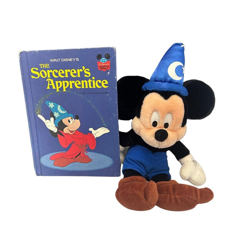 May include: A plush Mickey Mouse toy wearing a blue sorcerer's hat and outfit, next to a vintage book titled "The Sorcerer's Apprentice." The book cover is blue with Mickey Mouse in a red robe. The toy has brown shoes and white gloves.