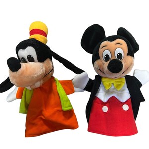 以下が含まれることがあります： ディズニーキャラクターのぬいぐるみハンドパペット2体。1つは、オレンジ色のシャツ、緑色のベスト、黄色の帽子をかぶったグーフィーです。もう1つは、赤いトップス、黒いジャケット、黄色の蝶ネクタイを着たミッキーマウスです。