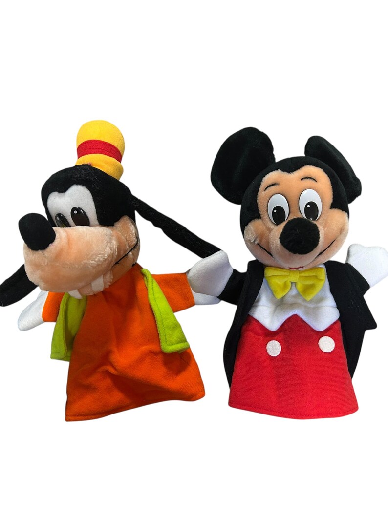以下が含まれることがあります： ディズニーキャラクターのぬいぐるみハンドパペット2体。1つは、オレンジ色のシャツ、緑色のベスト、黄色の帽子をかぶったグーフィーです。もう1つは、赤いトップス、黒いジャケット、黄色の蝶ネクタイを着たミッキーマウスです。