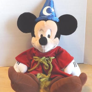 May include: A plush Mickey Mouse dressed as a sorcerer, wearing a blue hat with white stars and a crescent moon. He has a red robe with a gold rope belt and brown shoes. The classic Disney character is sitting down.