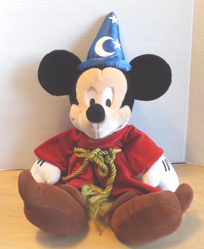 May include: A plush Mickey Mouse dressed as a sorcerer, wearing a blue hat with white stars and a crescent moon. He has a red robe with a gold rope belt and brown shoes. The classic Disney character is sitting down.