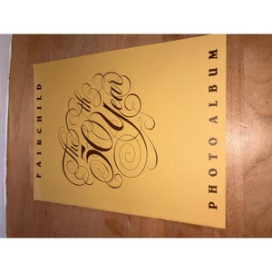 Puede incluir: Una cubierta de álbum de fotos rectangular de color amarillo con texto en marrón oscuro. El lado izquierdo dice FAIRCHILD verticalmente, el derecho dice PHOTO ALBUM verticalmente y el centro presenta texto en cursiva The 50th Year.
