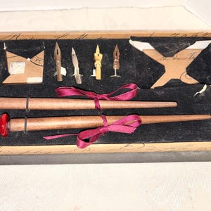 Può includere: Un set di calligrafia vintage in una scatola di legno con interno in velluto nero. Il set include portapenne in legno, vari pennini e un nastro rosso. I portapenne sono marroni con punta in vetro rosso. La scatola è decorata con scritte a mano.