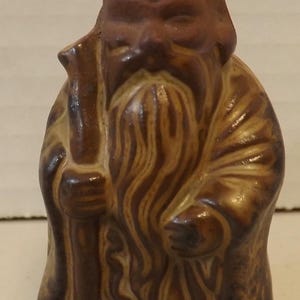 May include: A ceramic figurine of a wise old man with a long beard, holding a staff. The figure is primarily brown with lighter brown details, and the beard has a detailed, flowing texture. The figurine is a decorative item.