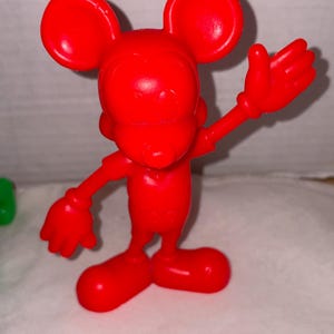 K&ouml;nnte beinhalten: Eine leuchtend rote Figur einer Zeichentrickmaus mit gro&szlig;en, runden Ohren und ausgestreckten Armen. Die Maus steht mit gespreizten F&uuml;&szlig;en da, und ihre rechte Hand ist zum Winken erhoben. Die Figur besteht aus einem glatten, gl&auml;nzenden Material.