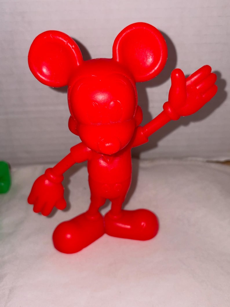 K&ouml;nnte beinhalten: Eine leuchtend rote Figur einer Zeichentrickmaus mit gro&szlig;en, runden Ohren und ausgestreckten Armen. Die Maus steht mit gespreizten F&uuml;&szlig;en da, und ihre rechte Hand ist zum Winken erhoben. Die Figur besteht aus einem glatten, gl&auml;nzenden Material.