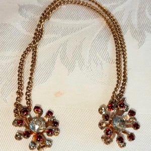 May include: A vintage gold-toned chain with two snowflake-shaped ornaments. Each ornament features a large clear crystal center surrounded by smaller red and clear crystals. The chain is draped on a white patterned surface.