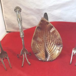 Può includere: Un assortimento di posate antiche su una superficie rossa. Il set include tre forchette d'argento e un piatto a forma di foglia con finitura strutturata marrone dorato. Le forchette hanno design diversi.