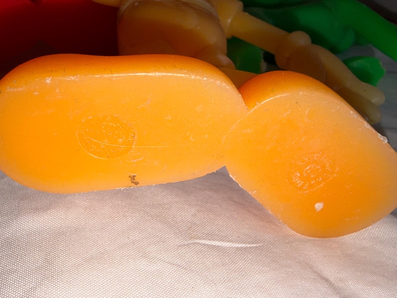 K&ouml;nnte beinhalten: Nahaufnahme von zwei orangefarbenen Spielzeugf&uuml;&szlig;en aus Kunststoff. Die F&uuml;&szlig;e haben eine leuchtende, einfarbige orange Farbe und scheinen Teil einer gr&ouml;&szlig;eren Spielfigur zu sein. Die F&uuml;&szlig;e liegen auf einer wei&szlig;en Oberfl&auml;che. Das Bild ist gut beleuchtet und hebt die glatte Textur des Kunststoffs hervor.