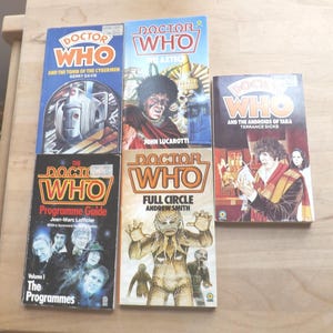 以下が含まれることがあります： ヴィンテージの「ドクター・フー」の本のコレクション。表紙には、サイバーマンやアステカ人など、シリーズのキャラクターやシーンのイラストが描かれています。本は明るい色の木製の表面に配置されています。
