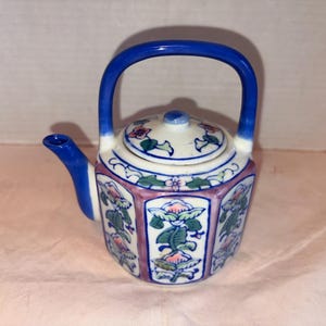 May include: A small, decorative ceramic teapot with a blue handle and spout. The teapot features a white lid and body, adorned with floral patterns in pink, green, and blue. The body has a hexagonal shape with pink borders.