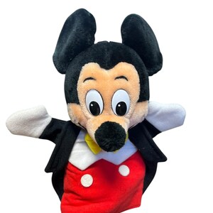以下が含まれることがあります： ミッキーマウスのハンドパペット。パペットは、大きな黒い耳、日焼けした顔、黒い鼻を持つ黒い頭をしています。黒いジャケット、白いボタンが付いた赤いシャツ、黄色の蝶ネクタイ、白い手袋を着用しています。