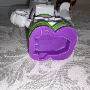 Puede incluir: Un juguete robot Buzz Lightyear blanco y morado, con detalles verdes y morados. El juguete est&aacute; tumbado de lado, con el pie morado visible. El juguete est&aacute; sobre una superficie blanca estampada.