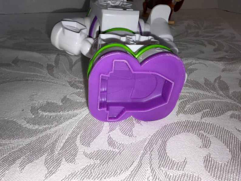Puede incluir: Un juguete robot Buzz Lightyear blanco y morado, con detalles verdes y morados. El juguete est&aacute; tumbado de lado, con el pie morado visible. El juguete est&aacute; sobre una superficie blanca estampada.