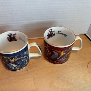 May include: Two ceramic mugs with white interiors and gold rims. One mug is navy blue with a floral design, the other is burgundy with a similar pattern. Both mugs have the word "Norway" printed on them.