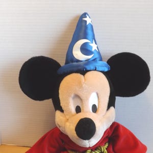 May include: A plush Mickey Mouse toy wearing a blue wizard hat with white stars and a crescent moon. Mickey has black ears, a beige face, and a red robe with a green and gold rope. The toy is a classic Disney character.