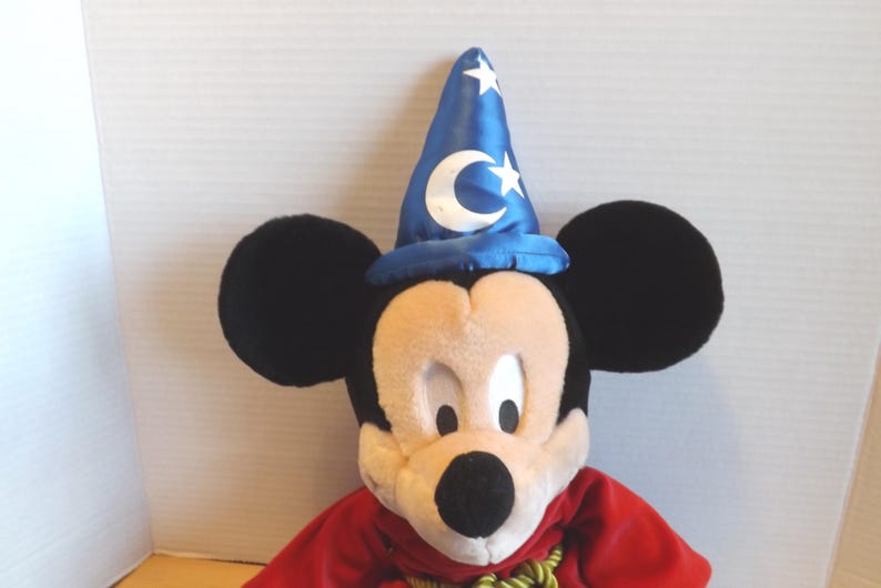 May include: A plush Mickey Mouse toy wearing a blue wizard hat with white stars and a crescent moon. Mickey has black ears, a beige face, and a red robe with a green and gold rope. The toy is a classic Disney character.