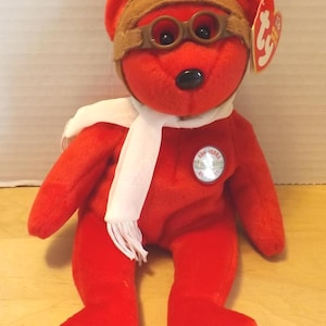 Puede incluir: Un oso de peluche rojo con gorro y gafas de aviador marrones, una bufanda blanca y un pin que dice "100 Years". El oso tiene una nariz negra y una etiqueta Ty.