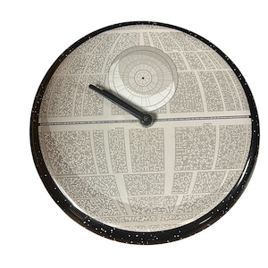 May include: A round wall clock featuring a detailed illustration of the Death Star from Star Wars. The clock face is primarily gray and white, with a black speckled border and black clock hands. The design is a detailed representation of the iconic space station.