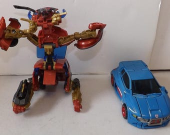 May include: A red, gold, and blue transforming robot toy in a partially assembled state, next to a blue toy car with red wheels. The robot has articulated limbs and a complex design, while the car features a sleek, modern aesthetic.