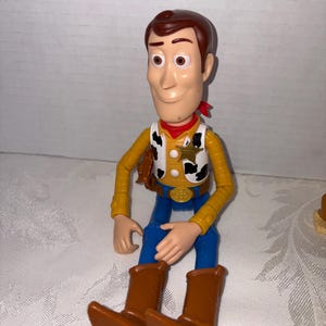 Puede incluir: Una figura de juguete de pl&aacute;stico del Sheriff Woody de la pel&iacute;cula animada Toy Story. La figura tiene un sombrero de vaquero marr&oacute;n, una camisa amarilla, pantalones azules y botas marrones. Una bandana roja est&aacute; alrededor del cuello. La figura est&aacute; sentada.