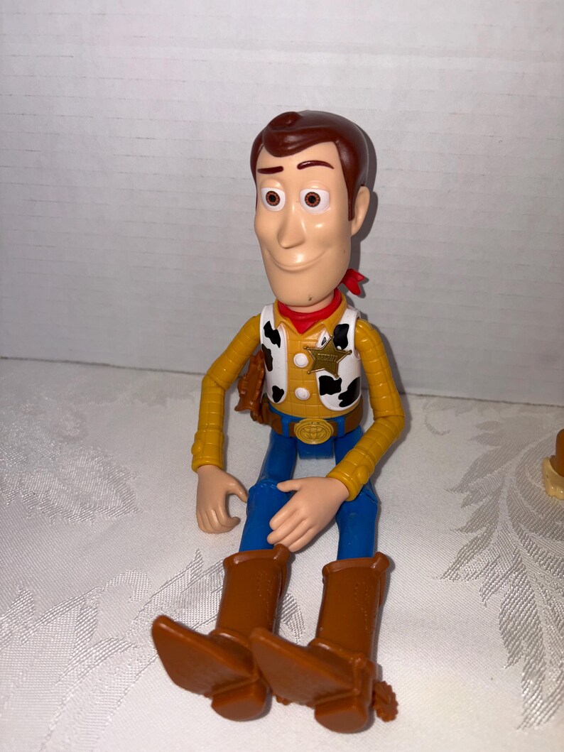 Puede incluir: Una figura de juguete de pl&aacute;stico del Sheriff Woody de la pel&iacute;cula animada Toy Story. La figura tiene un sombrero de vaquero marr&oacute;n, una camisa amarilla, pantalones azules y botas marrones. Una bandana roja est&aacute; alrededor del cuello. La figura est&aacute; sentada.