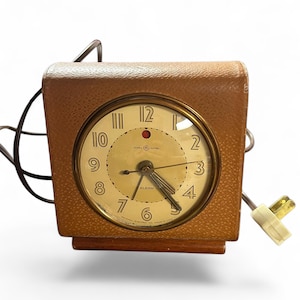 Puede incluir: Reloj despertador General Electric vintage con una caja marrón texturizada y una esfera redonda con borde dorado. La esfera del reloj es de color crema con números y manecillas negras. La palabra "ALARM" está impresa en la esfera. Un cable de alimentación y un enchufe están adjuntos.