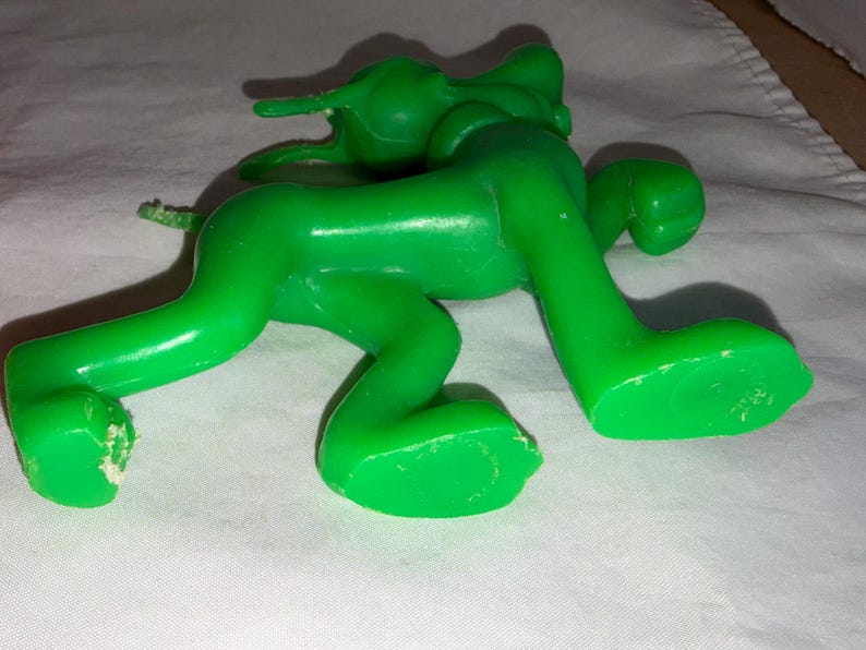 K&ouml;nnte beinhalten: Eine hellgr&uuml;ne Kunststoff-Spielfigur in Krabbelstellung. Das Spielzeug hat eine glatte, gl&auml;nzende Oberfl&auml;che und liegt auf einer wei&szlig;en Oberfl&auml;che. Die Figur hat einen langen Schwanz und gro&szlig;e F&uuml;&szlig;e. Das Spielzeug scheint ein Vintage- oder Retro-Artikel zu sein.