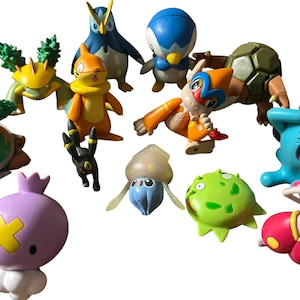 May include: A collection of various small plastic character figurines featuring diverse shapes and colors, including a purple round figure with a yellow cross, a blue creature with a wide grin, an orange bipedal character, and several green and brown creatures.