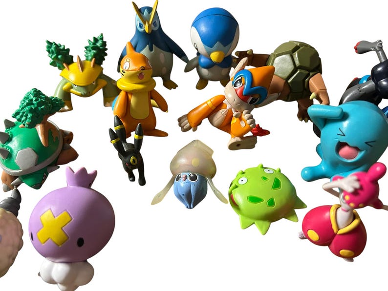 May include: A collection of various small plastic character figurines featuring diverse shapes and colors, including a purple round figure with a yellow cross, a blue creature with a wide grin, an orange bipedal character, and several green and brown creatures.