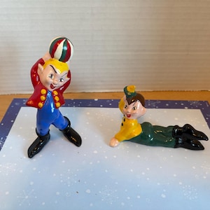 Pode incluir: Duas figuras de cerâmica de artistas de circo. Uma figura está em pé, usando um casaco vermelho, calças azuis e botas pretas, e segurando uma bola listrada. A outra figura está deitada, usando uma camisa amarela e calças verdes.