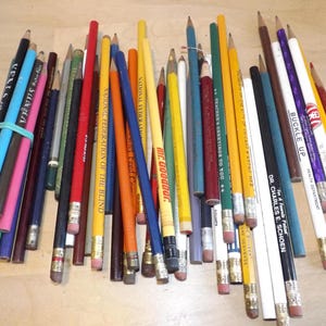 Puede incluir: Una colección de lápices de varios colores, incluyendo azul, amarillo, rojo y verde. Algunos lápices tienen texto impreso, como "VENUS" y "BUCKLE UP". Los lápices están dispuestos sobre una superficie de madera clara.