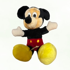 Puede incluir: Un peluche de Mickey Mouse con cuerpo negro, pantalones cortos rojos y zapatos amarillos. El juguete tiene una cara beige, orejas negras y guantes blancos. El personaje de Disney est&aacute; sentado con los brazos extendidos.