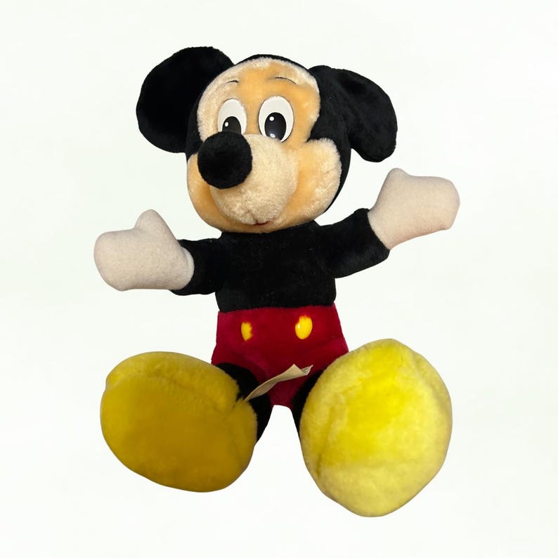 Puede incluir: Un peluche de Mickey Mouse con cuerpo negro, pantalones cortos rojos y zapatos amarillos. El juguete tiene una cara beige, orejas negras y guantes blancos. El personaje de Disney est&aacute; sentado con los brazos extendidos.