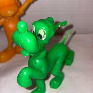 K&ouml;nnte beinhalten: Eine gr&uuml;ne Cartoon-Hundefigur aus Kunststoff mit wei&szlig;en Augen und einem kleinen Schwanz. Der Hund befindet sich in einer Laufhaltung. Im Hintergrund befindet sich eine orangefarbene Cartoonfigur aus Kunststoff.