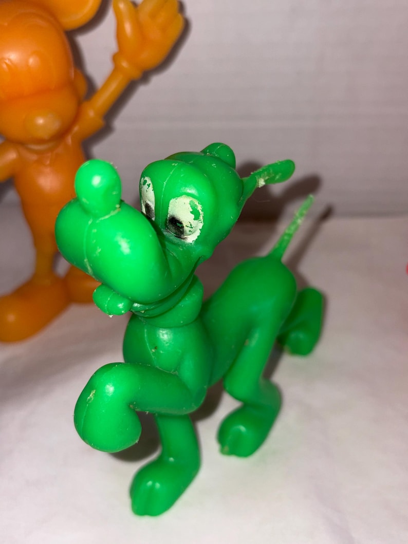 K&ouml;nnte beinhalten: Eine gr&uuml;ne Cartoon-Hundefigur aus Kunststoff mit wei&szlig;en Augen und einem kleinen Schwanz. Der Hund befindet sich in einer Laufhaltung. Im Hintergrund befindet sich eine orangefarbene Cartoonfigur aus Kunststoff.