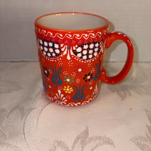 May include: A ceramic mug with a vibrant orange base color and a white interior. The mug is adorned with intricate floral and geometric patterns in blue, white, and yellow. It has a rounded handle and a decorative rim.