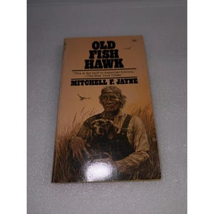 May include: A paperback book titled Old Fish Hawk by Mitchell F. Jayne with a tan cover. The cover features an illustration of a person with a dog in a field of tall grass, with a small bird flying above and the text This is the stuff of American folklore.