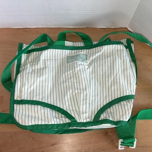 May include: A white and green striped backpack with green trim and straps. The backpack has a top handle and adjustable shoulder straps. The bag has a rectangular shape with rounded corners and a white plastic buckle.