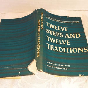 以下が含まれることがあります： アルコホーリクス・アノニマスの「Twelve Steps and Twelve Traditions」というタイトルの開いた本。表紙はティール色で、黒い横線があり、タイトルは大きな金色の文字で書かれています。本は少し傷んでいます。