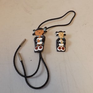 Puede incluir: Un conjunto de corbata bolo y colgante con figuras de estilo nativo americano. Las figuras están pintadas con piel morena, cabello negro y vestimenta tradicional. La corbata bolo tiene un cordón trenzado negro con puntas plateadas.
