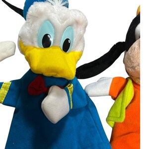以下が含まれることがあります： 2つのぬいぐるみキャラクターの人形をクローズアップ。1つはドナルドダックで、白い頭、青い帽子、黄色のくちばし、青い衣装です。もう1つはグーフィーで、オレンジ色の衣装と緑のベストを着ています。どちらも黒い目です。