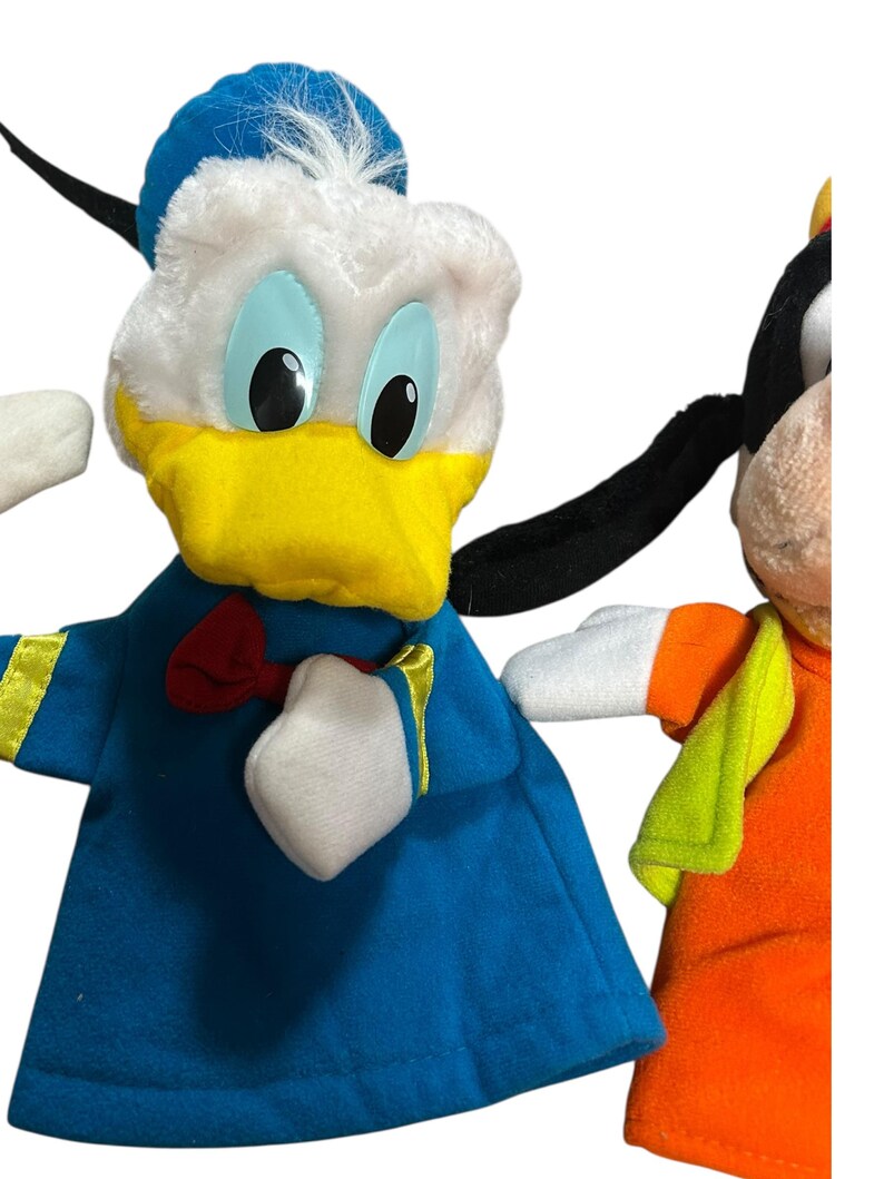 以下が含まれることがあります： 2つのぬいぐるみキャラクターの人形をクローズアップ。1つはドナルドダックで、白い頭、青い帽子、黄色のくちばし、青い衣装です。もう1つはグーフィーで、オレンジ色の衣装と緑のベストを着ています。どちらも黒い目です。