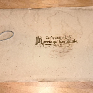 May include: Vintage marriage certificate with the text "Our Voyage of Life Marriage Certificate" in ornate lettering. The aged paper has water stains and is tied with a gray string. The certificate is a cream color.