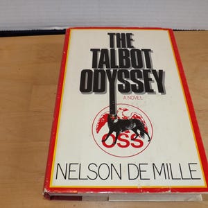 Könnte beinhalten: Ein Buch mit dem Titel "The Talbot Odyssey: A Novel" von Nelson DeMille. Das Cover ist cremefarben mit schwarzem Text und einem rot-schwarzen Emblem. Das Buch hat einen rot-gelben Rand.
