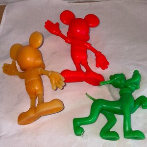K&ouml;nnte beinhalten: Drei Vintage-Figuren von Disney-Charakteren: eine rote Mickey Mouse, eine orangefarbene Mickey Mouse und ein gr&uuml;ner Pluto. Die Mickey Mouse-Figuren stehen mit ausgestreckten Armen. Die Pluto-Figur befindet sich in einer Laufhaltung.